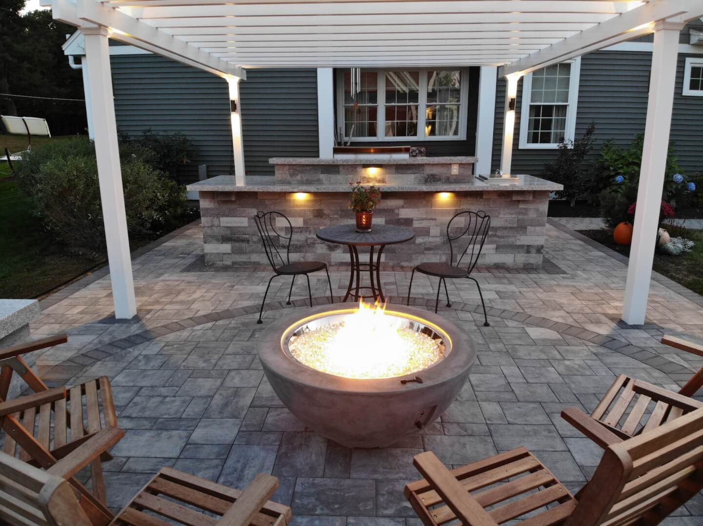 Outdoor fire pit area with wooden chairs, stone seating wall, pergola, and built-in bar on a custom paver patio
