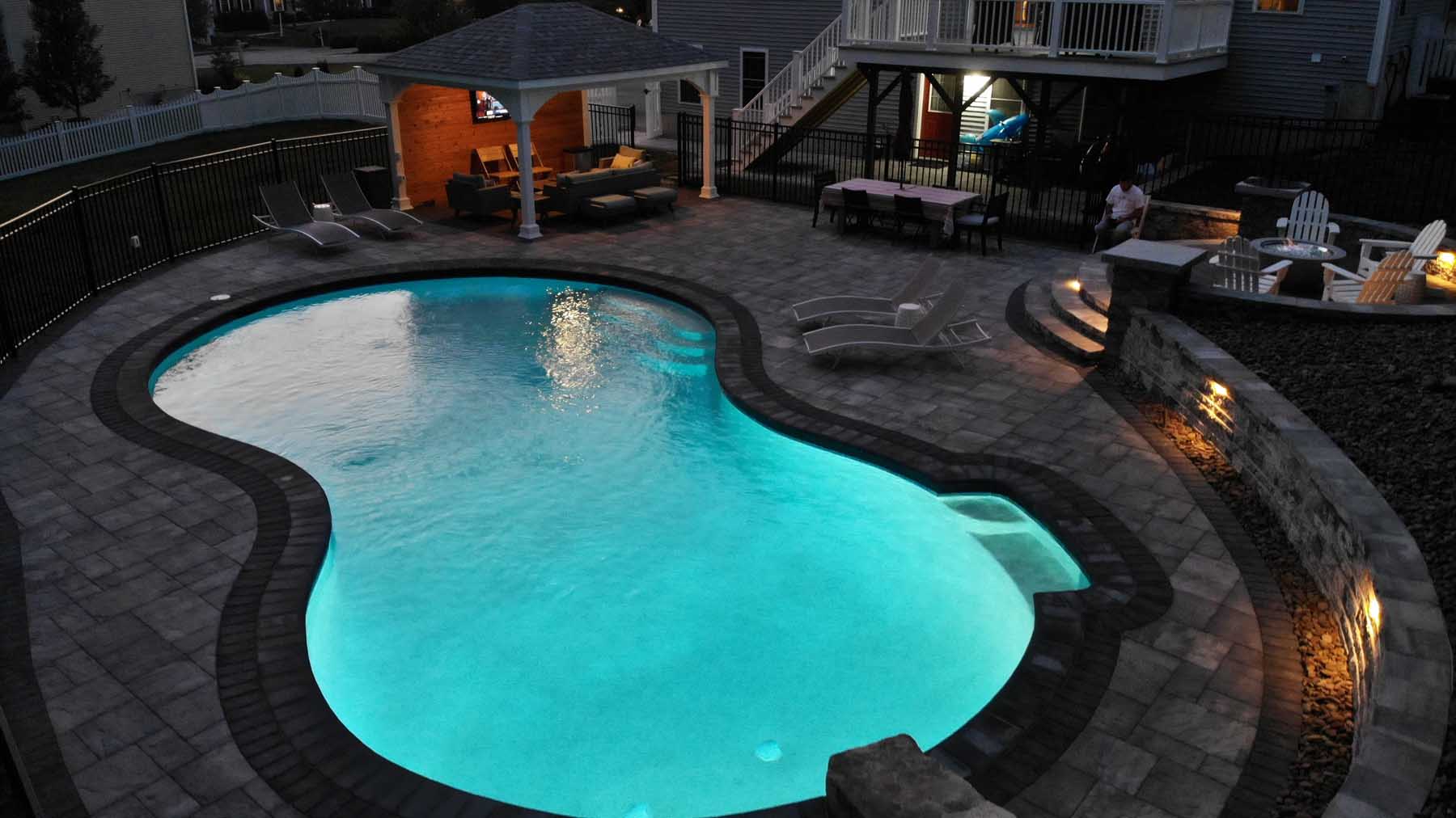 Evening view of a glowing in-ground pool with custom paver deck, built-in lighting, covered cabana with outdoor seating, and fire pit lounge area