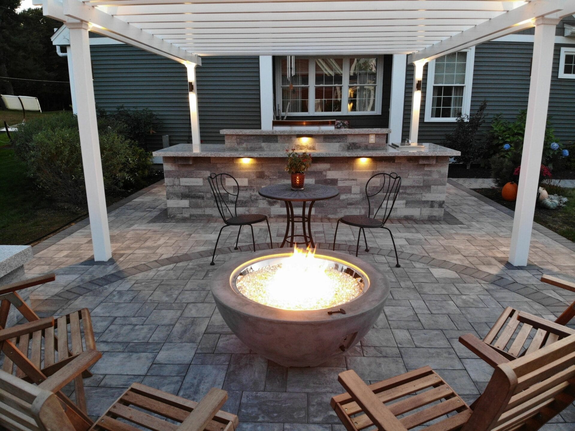 Backyard patio with a fire pit, wooden chairs, pergola, and an outdoor kitchen with built-in lighting