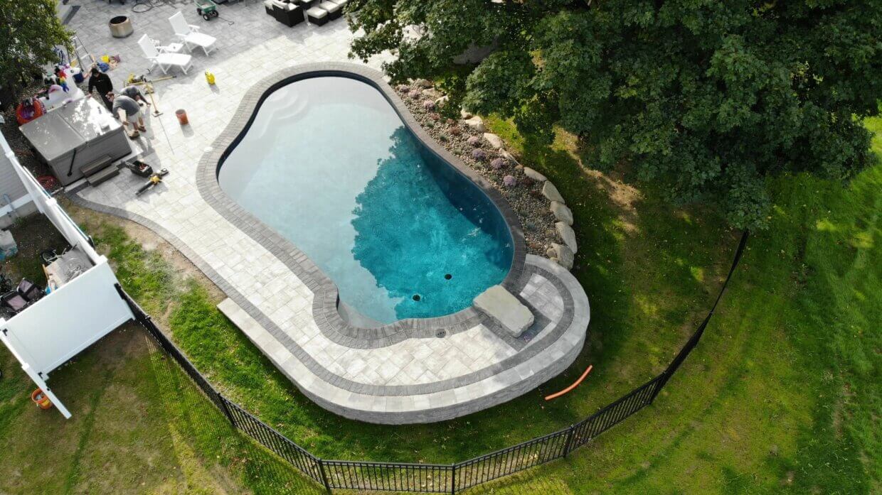 Aerial view of a kidney-shaped in-ground pool with light stone paver deck, hot tub installation, and landscaping along one side, surrounded by a black fence