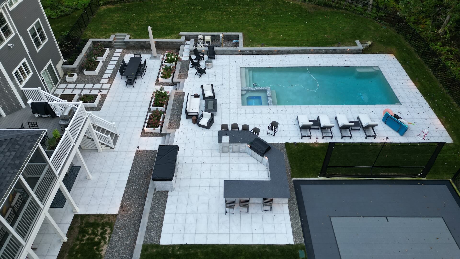 Overhead view of a luxury backyard with white tile patio, outdoor kitchen, lounge seating, in-ground pool, and modern landscaping features