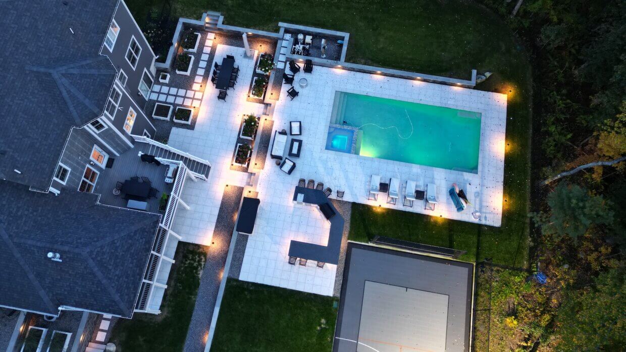 Aerial view of a luxury backyard with a modern rectangular pool, white paver patio, multiple lounge areas, and custom lighting surrounding a large home