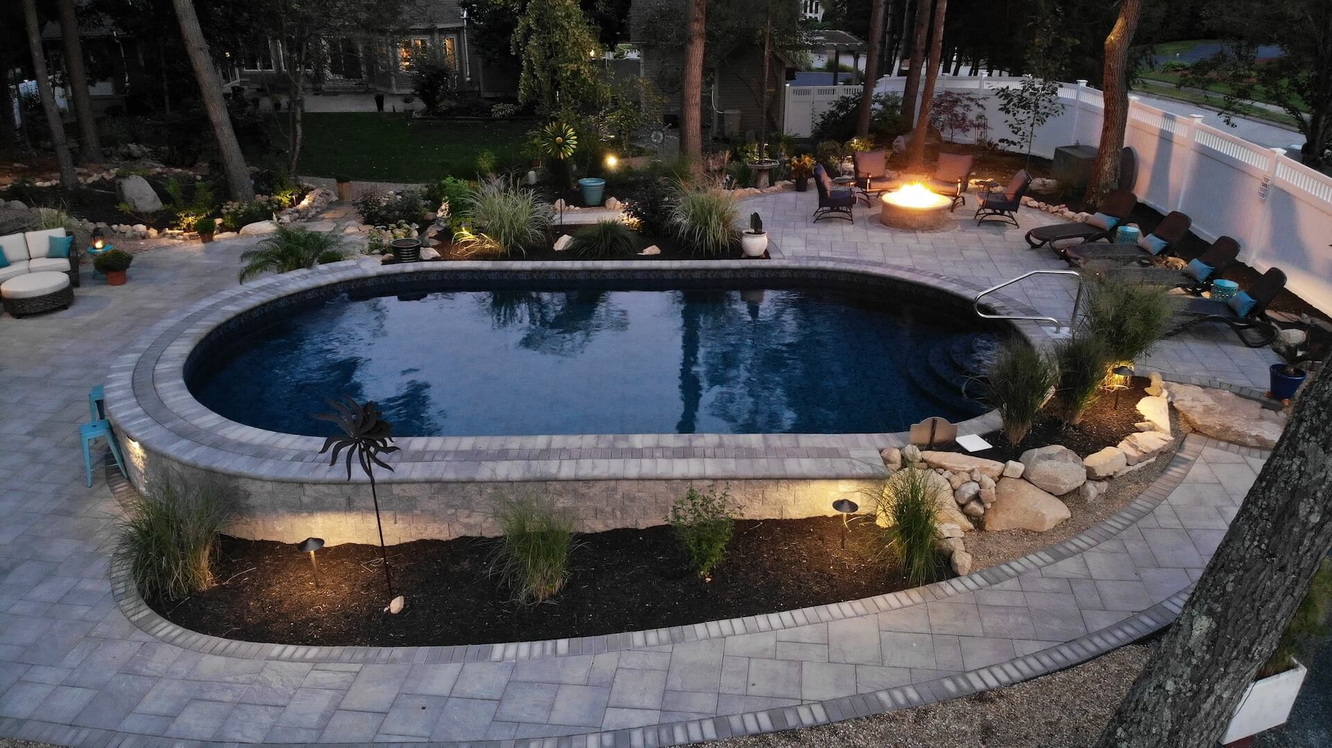 Backyard poolscape with custom stone pavers, fire pit seating area, lounge chairs, and landscape lighting creating a serene evening ambiance