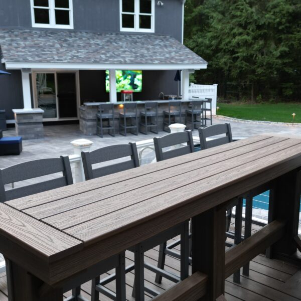 View from a raised deck showing a high-top outdoor dining table and chairs overlooking a backyard patio with lounge seating, a covered outdoor bar, and a swimming pool beside a gray-sided home.