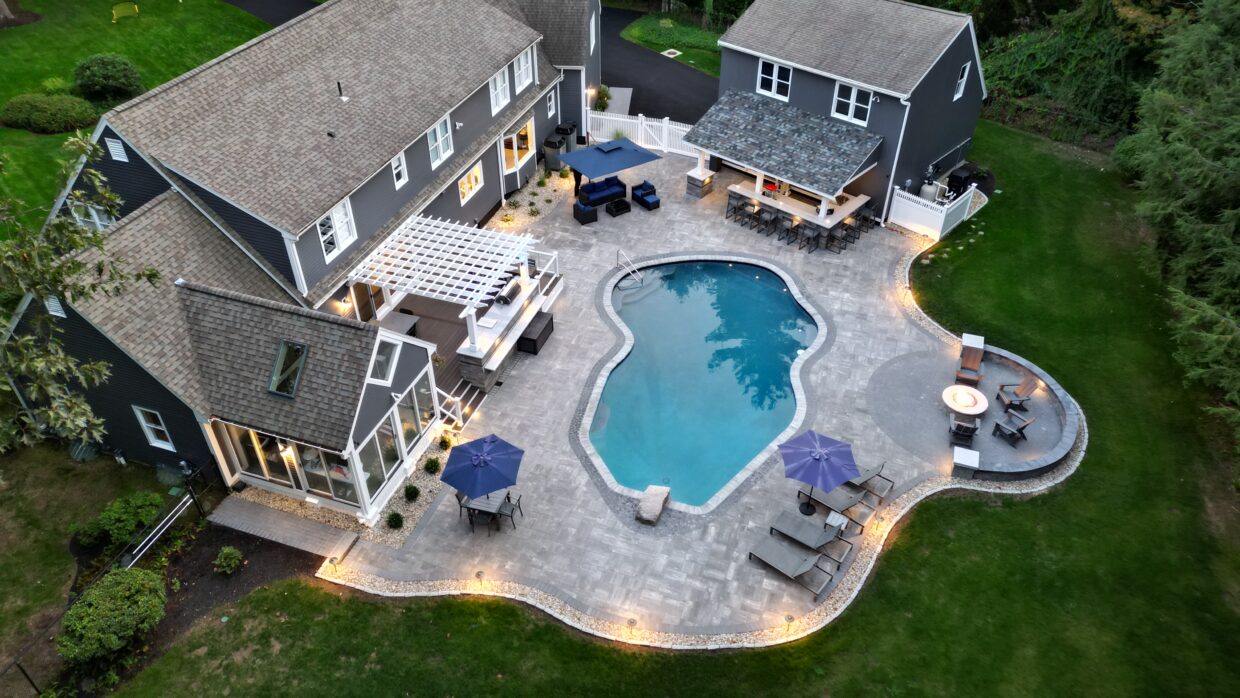 Aerial view of a large backyard outdoor living area featuring a freeform swimming pool, expansive stone paver patio, pergola-covered deck, outdoor bar pavilion, fire pit seating area, and multiple dining and lounge spaces surrounding a gray two-story home.