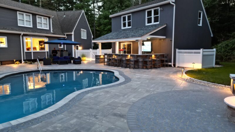 Evening view of a backyard outdoor living space with a curved swimming pool, stone patio, circular fire pit with Adirondack chairs, a covered outdoor bar, and gray-sided buildings surrounded by trees.