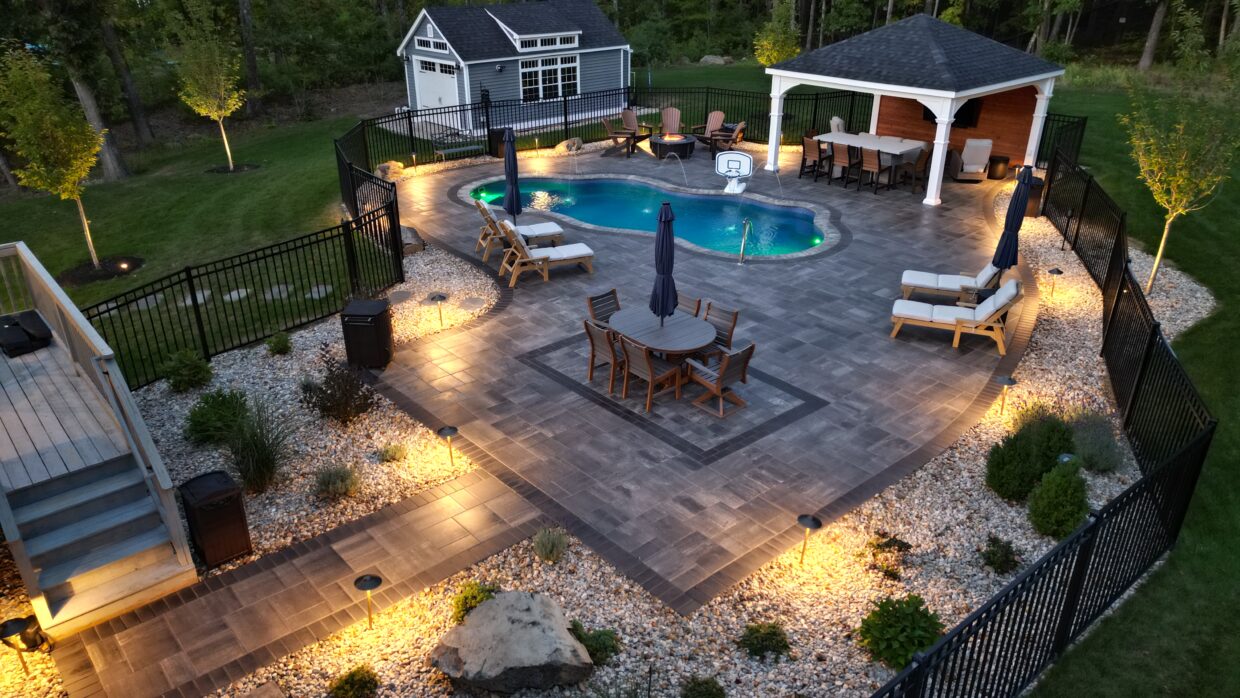 Aerial view of a landscaped backyard with a freeform swimming pool, expansive stone patio, dining and lounge areas, a covered pavilion, fire pit seating, and pathway lighting enclosed by a black metal fence at dusk.