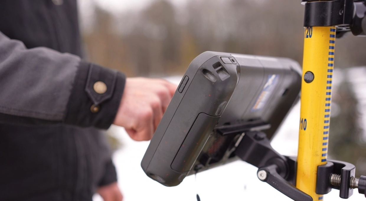 close-up of a field technician using a handheld data collection device mounted on a survey rod for measuring and recording site elevations outdoors